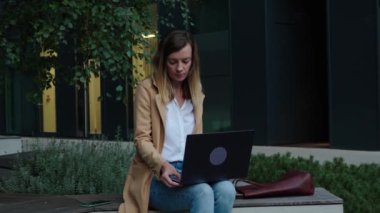 Uzaktan çalışan bir kadın, şehir caddesinde bankta oturuyor ve dizüstü bilgisayar kullanıyor. Çevrimiçi serbest çalışan, dizüstü bilgisayarlı klavyede yazan bir kadın.
