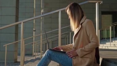 Uzaktan çalışan bir kadın, şehir caddesindeki merdivenlerde oturuyor ve dizüstü bilgisayar kullanıyor. Çevrimiçi serbest çalışan, dizüstü bilgisayarlı klavyede yazan bir kadın.