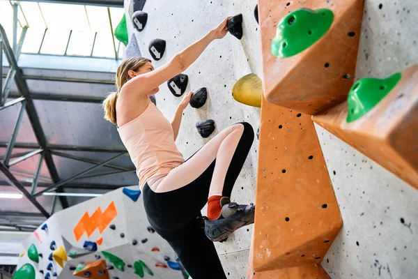Woman climbing up on wall at bouldering gym. Female climber training, hanging on bouldering climbing wall. Active lifestyle and extreme sport concept.