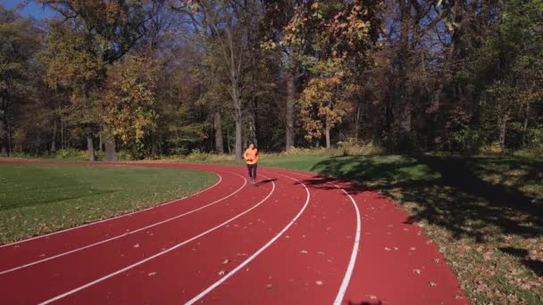 身穿橙色运动服的男子在跑道上奔跑 空中无人驾驶飞机的视野 男子跑步运动员在体育场慢跑 秋季在室外进行积极的体育锻炼 健康的生活方式 — 图库视频影像