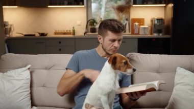 Oturma odasında kanepede oturan, kağıt kitap okuyan ve köpeği okşayan bir adam. Evde evcil hayvanla dinlenmek, kitapla rahatlamak