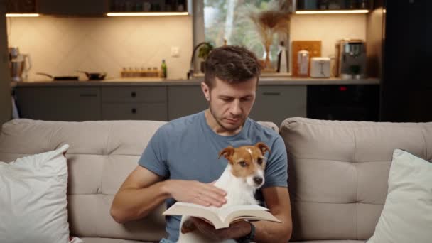 Mann Sitzt Auf Sofa Wohnzimmer Liest Papierbuch Und Streichelt Hund — Stockvideo