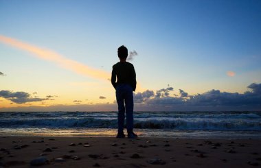 Boy silhouette standing on beach near sea and looking on the waves in sunset