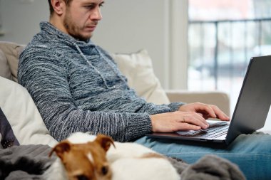 Man sitting on sofa in living room and using laptop, near lying dog. Freelancer working at home workplace. Remote work and work from home concept