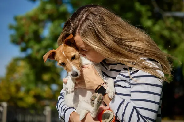 Woman holds her dog on hands and embracing with it. Happy female owner hugging and kissing cute friend. Pet care