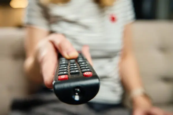 Woman holds TV remote control. Female is sitting on sofa in living room and switching channels on the TV. Concept of entertainment and lazy weekends
