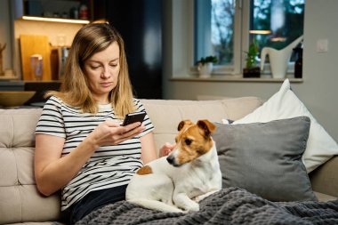Evde cep telefonu kullanan bir kadın. Kanepede köpeğiyle oturan ve akıllı telefondan sosyal ağlara bakan bir kadın. Cep telefonundan online alışveriş.