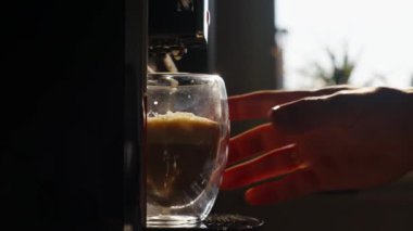 Otomatik kahve makinesi sabah güneşiyle birlikte bardakta kapuçino hazırlıyor, erkek eli bir fincan kahve alıyor. Günlük sabah rutini
