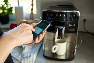 Kahve makinesini çalıştırmak için akıllı telefon uygulaması kullanıyor. Kadın, modern espresso makinesiyle taze kahve hazırlıyor.