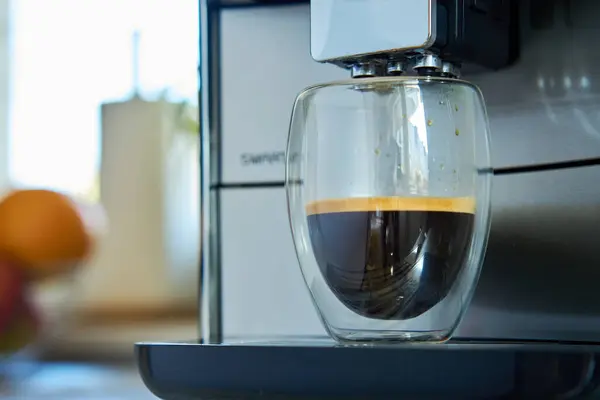 Fresh espresso in clear glass at morning. Coffee machine in kitchen, close up. Modern coffee maker with freshly brewed coffee in glass cup. Kitchen appliances
