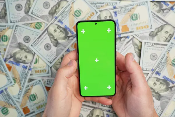 Hands holding smartphone with green screen on pile of dollar banknotes, top view. Concept of online earnings