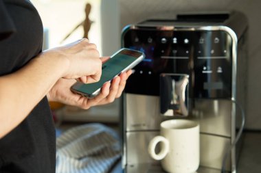 Kahve makinesini çalıştırmak için akıllı telefon uygulaması kullanıyor. Kadın, modern espresso makinesiyle taze kahve hazırlıyor.