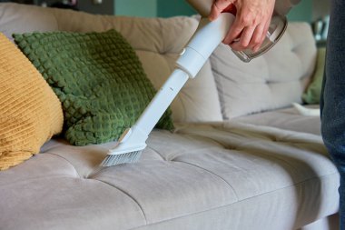 Kadın oturma odasındaki kanepenin yüzeyini temizlemek için portatif kablosuz elektrik süpürgesi kullanıyor. Ev temizliği ve el süpürgesi. Ev işleri.