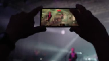 Sahnenin önünde duran kişi, akıllı telefondan canlı müzik konseri kaydediyor. Sahneye yakın insanlar müzisyen gruplarının performansından hoşlanırlar