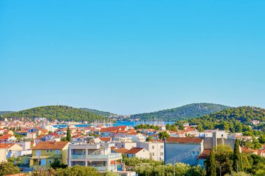 Kırmızı çatılı evleri ve açık mavi gökyüzünün altında yemyeşil tepeleri olan kıyı kasabası manzarası. Yaz tatilleri için Adriyatik Denizi yakınlarındaki Akdeniz turistik kasabası