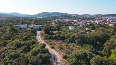 Aerial view of man jogging on rocky trail surrounded by lush greenery and trees in coastal town with sea landscape. Male sportsman running cross country on hot summer day. Endurance training