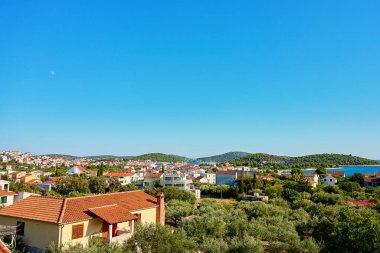 Kırmızı çatılı evleri ve açık mavi gökyüzünün altında yemyeşil tepeleri olan kıyı kasabası manzarası. Yaz tatilleri için Adriyatik Denizi yakınlarındaki Akdeniz turistik kasabası