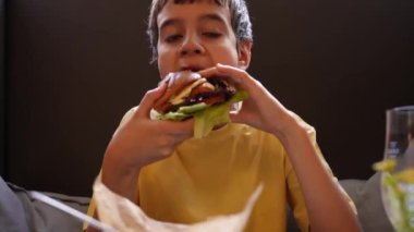 Restoranda salatalı, turşulu ve domatesli tavuk burger yiyen çocuk. Çocuk hamburgeri ısırırken yakın çekim. Aç çocuk sağlıksız yiyecekler yiyor.