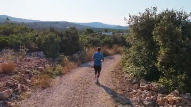 Yeşillik ve açık mavi gökyüzünün altındaki ağaçlarla çevrili kayalık yolda koşan bir adam. Sıcak yaz gününde ülkeyi boydan boya koşan erkek sporcu. Dayanıklılık eğitimi