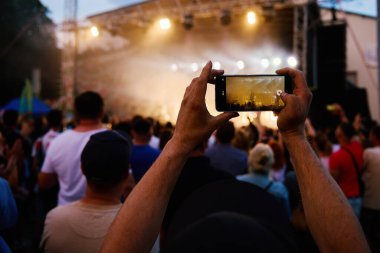 Sahnenin önünde duran kişi, akıllı telefondan canlı müzik konseri kaydediyor. Sahneye yakın insanlar müzisyen gruplarının performansından hoşlanırlar