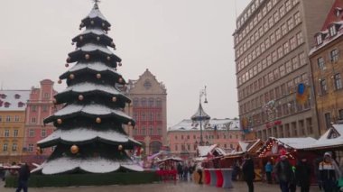 Wroclaw, Polonya 'da Noel pazarı. Karla kaplı büyük bir Noel ağacı. İnsanlar şehir caddesinde şenlikli süslemelerle yürüyor. Wroclaw, Polonya - 6 Aralık 2023