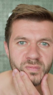Banyoda traşsız yüzlere dokunan bir adamın aynadaki sakal uzamasını incelerken. Cilt bakımı ve kişisel bakım konsepti