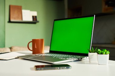 Yeşil ekran, akıllı telefon, kahve fincanı ve küçük saksı bitkilerinin yer aldığı modern ev çalışma alanı. Mutfağı arka planda olan serbest çalışanlar için merkez ofis. Üretkenlik ve uzak çalışma kavramı