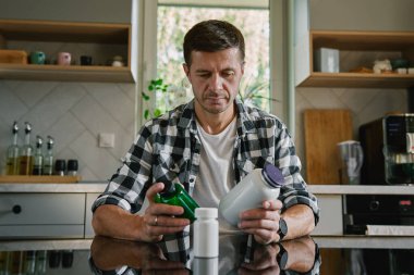 Adam mutfakta masada oturmuş çeşitli ilaç kapsülleri şişelerine bakıyor. İnsan ilaç ya da vitamin alıyor. İlave, ilaç veya sağlık hizmetleri kavramı