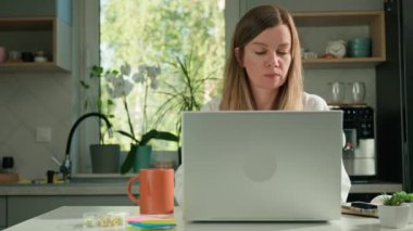 Serbest çalışan kadın, iş gününü mutfaktaki dizüstü bilgisayarını kapatarak tamamlıyor. Kadın, ofisteki masasında dizüstü bilgisayar ve kırtasiyeyle işini bitiriyor.