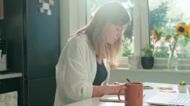 Kadın ofis masasında uzaktan çalışıyor, not defterine not alıyor. Kadın serbest çalışanın sabahları dizüstü bilgisayar kullanışının arka görüntüsü