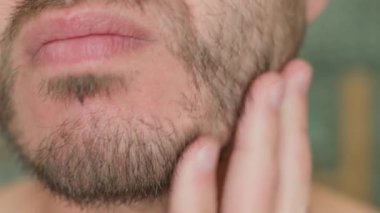 Banyoda traşsız yüzlere dokunan bir adamın aynadaki sakal uzamasını incelerken. Cilt bakımı ve kişisel bakım konsepti