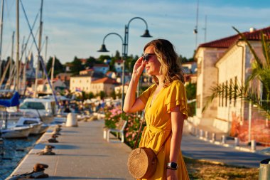 Kadın, palmiye ağaçları ve çiçeklerle güneşli güvertede tek başına yürüyor. Kadın turist, sahil kasabasında gün batımında marina boyunca geziniyor.