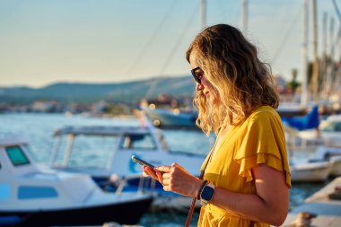 Sarı elbiseli genç kadın, sahil kasabasında güneşli şehir caddesinde yürürken akıllı telefonunu kullanıyor. Kadın turist gün batımında marinada dururken akıllı telefonunu kontrol ediyor.