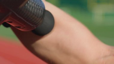 Stadyum pistindeki antrenmandan sonra iyileşmek için kol kaslarına elle masaj tabancası kullanan erkek atletlerin yakın çekimi. Kas gevşemesi ve spor iyileştirmesi kavramı