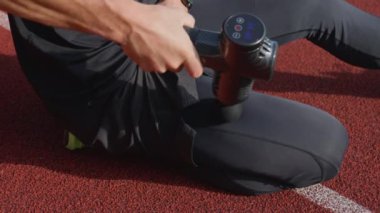 Stadyum pistindeki antrenmandan sonra iyileşmek için el bilgisayarı kullanarak bacak kaslarına masaj yapan erkek sporcunun yakın çekimi. Kas gevşemesi ve spor iyileştirmesi kavramı