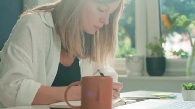 Masada dizüstü bilgisayar kullanan ve deftere yazı yazan serbest çalışan bir kadın. Ev ofisinde uzaktan çalışma