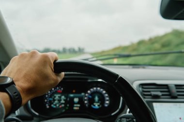 Sürücü direksiyonu sıkıca kavrıyor. İlerideki kırsal yol ön camdan görünüyor. Yağmurlu bir günde araba süren bir adam. Modern aracın iç görünümü