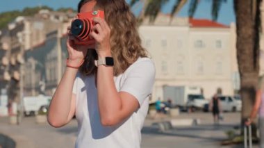 Kadın, arka planda palmiye ağaçları ve tarihi binalarla kıyı kasabasında fotoğraf çekiyor. Kadın turist, seyahat sırasında eski moda kamera ile anıları yakalıyor.