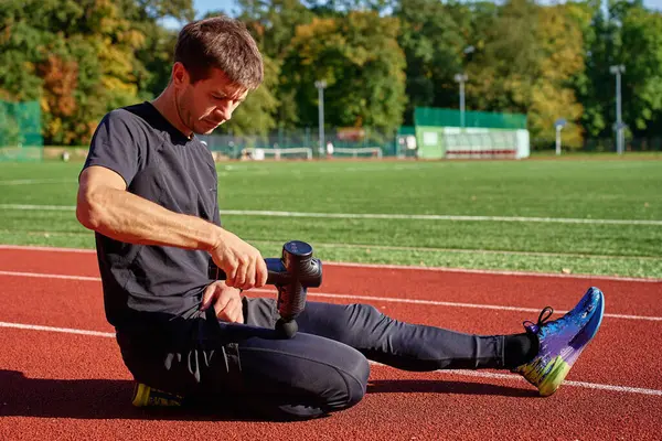 Stadyum pistindeki antrenmandan sonra iyileşmek için el bilgisayarı kullanarak bacak kaslarına masaj yapan erkek sporcunun yakın çekimi. Kas gevşemesi ve spor iyileştirmesi kavramı
