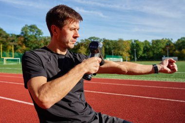 Stadyum pistindeki antrenmandan sonra iyileşmek için kol kaslarına elle masaj tabancası kullanan erkek atletlerin yakın çekimi. Kas gevşemesi ve spor iyileştirmesi kavramı