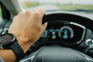 Sürücü direksiyonu sıkıca kavrıyor. İlerideki kırsal yol ön camdan görünüyor. Yağmurlu bir günde araba süren bir adam. Modern aracın iç görünümü