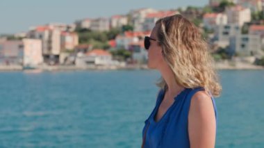 Mavi elbiseli ve güneş gözlüklü kadın yaz tatilinin keyfini çıkarıyor, Hırvatistan 'ın Dalmaçya kıyı şeridinde arka planda manzaralı bir kasabayla birlikte yürüyor.