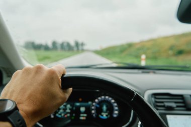 Driver hand gripping steering wheel with view of countryside road ahead through windshield. Man driving car in rainy day. Inside view of modern vehicle interior clipart