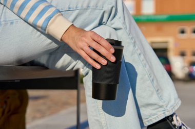 Woman in striped sweater and blue jeans enjoying her coffee break outdoors, holding black reusable coffee cup clipart