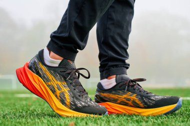 Yapay çimlerin üzerinde renkli Asics Novablast giyen yakın plan bir sporcu. Spor ayakkabılarıyla yeşil çimlerin üzerinde sporcu bacakları. Wroclaw, Polonya - 6 Kasım 2024