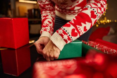 Noel hediyelerini kırmızı ve yeşil kağıda saran kadın elleri. Şenlikli kazaklı kadın kış tatili için hediyeler hazırlıyor. Noel ve Yeni Yıl Aile Gelenekleri