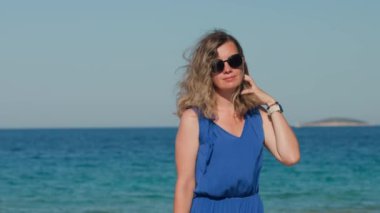Mavi elbiseli ve güneş gözlüklü rüzgârlı bir günde uçan saçlı bir kadın deniz kıyısında duruyor. Güneşli sahil boyunca yürüyen kadın turist. Yaz seyahati