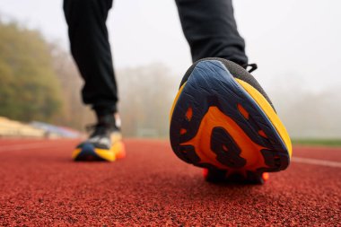 Kırmızı pist yüzeyinde spor yapmaya hazır renkli koşu ayakkabıları giyen bir sporcunun ayaklarının yakın çekimi. Spor ayakkabılı sporcu bacakları, yaklaşın. Fitness ve sağlıklı yaşam tarzı