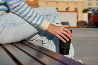 Woman in striped sweater and blue jeans enjoying her coffee break outdoors, holding black reusable coffee cup clipart