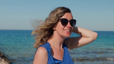Mavi elbiseli ve güneş gözlüklü rüzgârlı bir günde uçan saçlı bir kadın deniz kıyısında duruyor. Güneşli sahil boyunca yürüyen kadın turist. Yaz seyahati